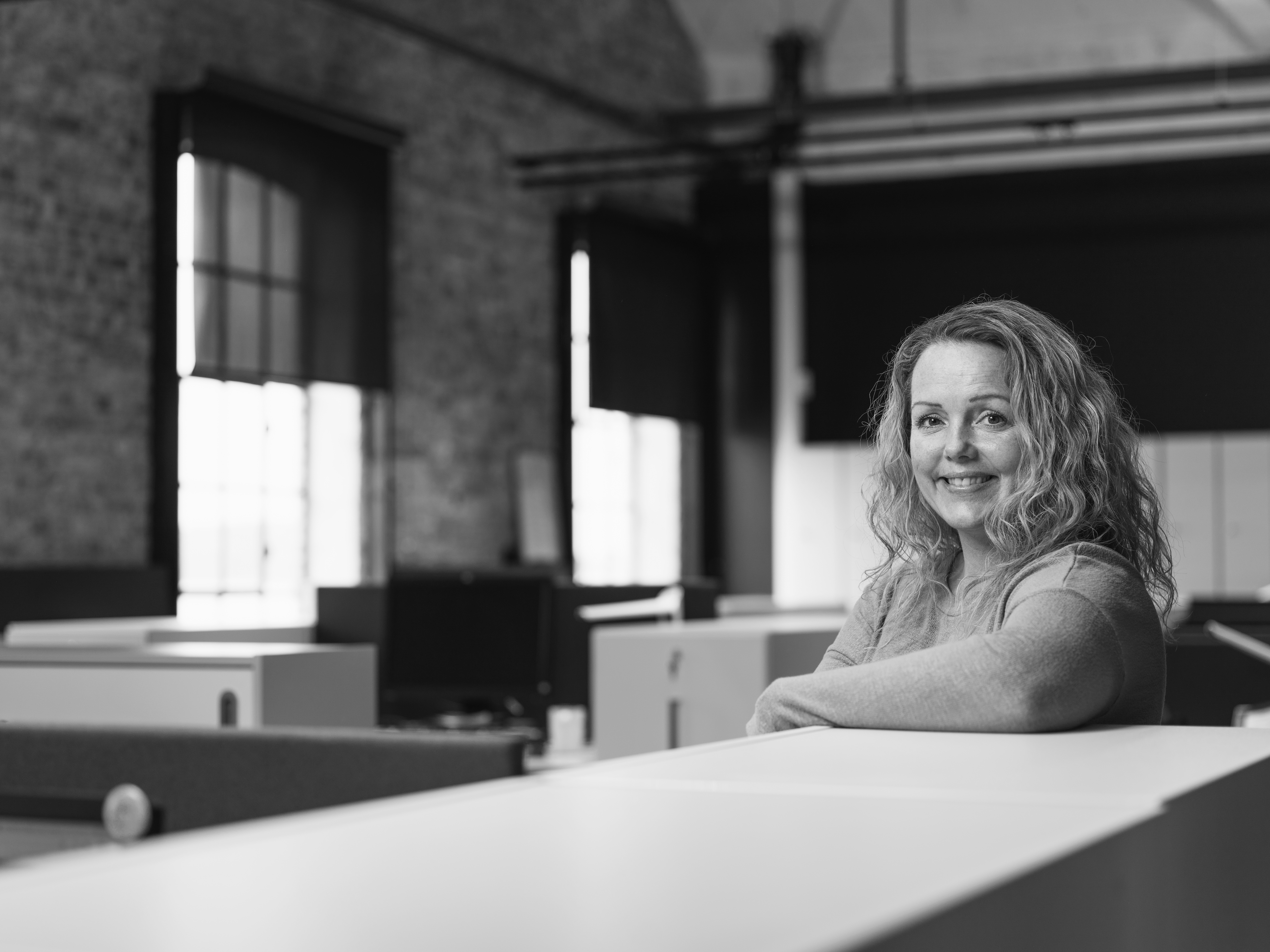 Kvinne som smiler i et moderne kontorlandskap hos Byggfakta, med store vinduer og murvegger i bakgrunnen, avbildet i svart-hvitt.
